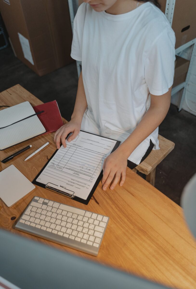 A woman reviews documents on a clipboard at a wooden desk, viewed from above.